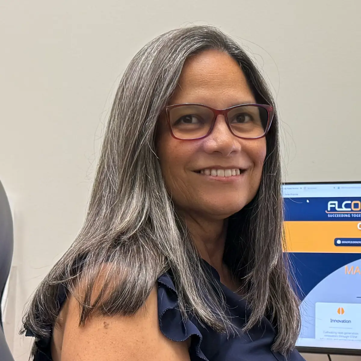 Smiling woman with shoulder-length gray hair and red glasses, standing beside a computer monitor in an office setting.
